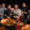 A group of musicians pose around a drumset