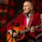 Gordon Lightfoot, in a deep red blazer, strums an acoustic guitar and sings into a mic