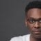Headshot of Shawn Okpebholo, looking straight at camera wearing black half-frame glasses and white t-shirt.,