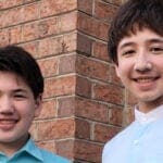 Brothers Joseph and Samuel Kim, both dressed in blue, pose together in front of a brick wall