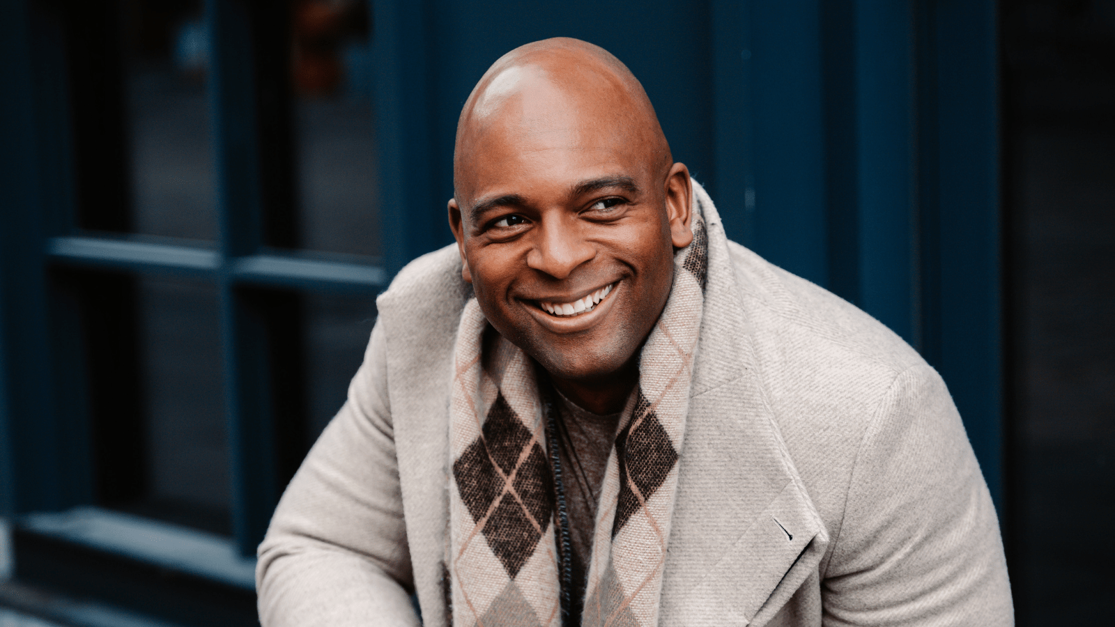 Portrait of Ryan Speedo Green in light wool coat and argyle scarf, dark background.