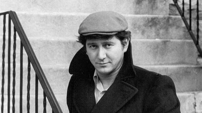 man in pea coat and snap brim hat looks at camera