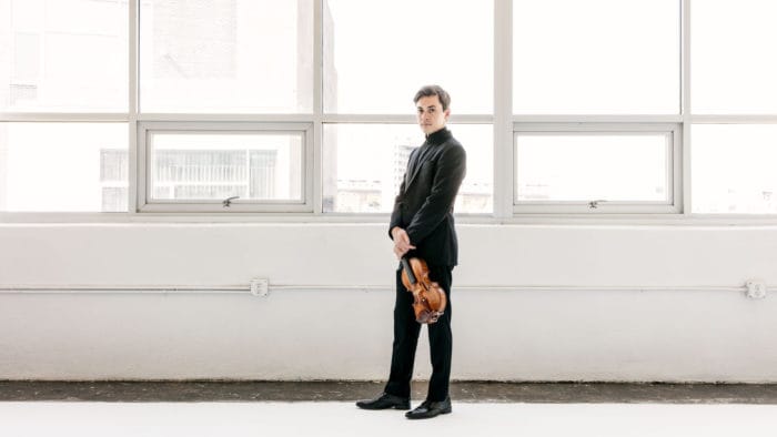 Benjamin Beilman poses with a violin in a brightly lit white industrial room