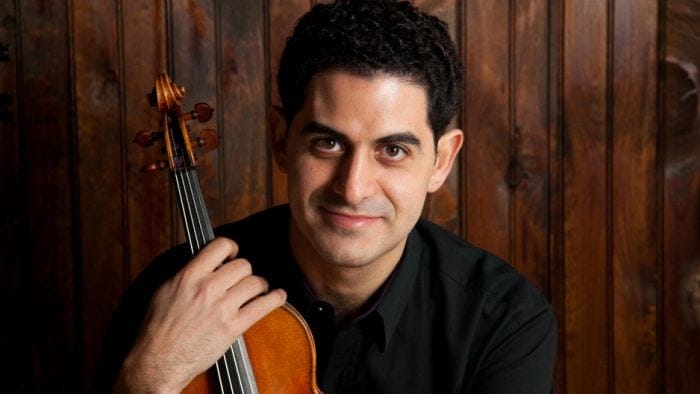 Violinist Arnaud Sussman holds a violin in front of a wood background