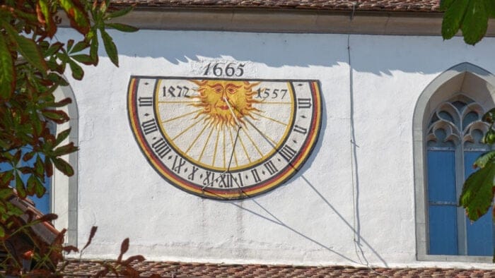 A sundial on the outside wall of a church. An anthropomorphic sun face's rays meet each of the numerals representing the hours.