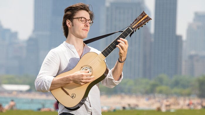Brandon Acker, plucking at a guitar, stands in the bright sunlight with the Chicago skyline in the background
