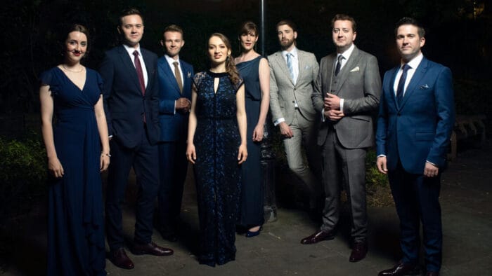 Portrait of eight singers of VOCES8 in mostly dark, semi-formal attire at night under a street lamp.