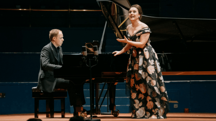 Louise Alder in dark floral gown, singing, gesturing towards audience with Joseph Middleton accompanying her at piano gown