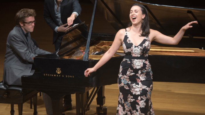 Kara Dugan in the crook of grand piano singing and gesturing joyfully in floral dress.