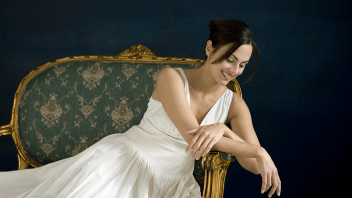 Isabel Leonard in white sun dress leaning over the arm of an antique upholstered chair, laughing
