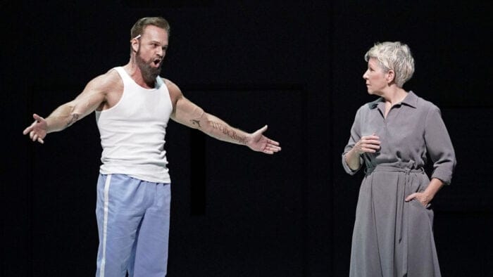 A scene from Dead Man Walking: Ryan McKinny, dressed as an inmate and with a cigarette tucked behind his ear, glowers with arms wide at a startled-looking Joyce DiDonato, wearing a gray dress