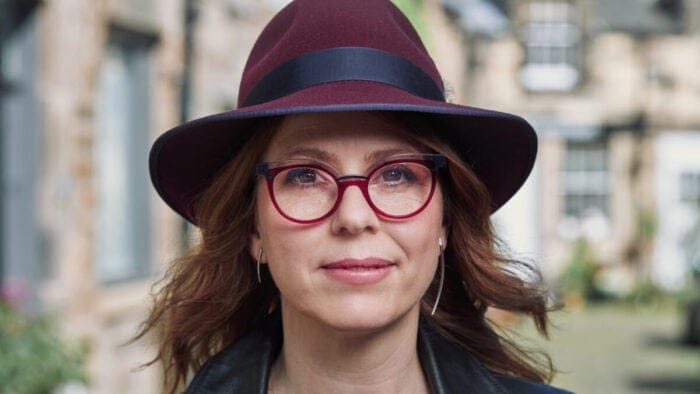 Anna Clyne, sporting a wine red fedora and glasses, stands in a courtyard