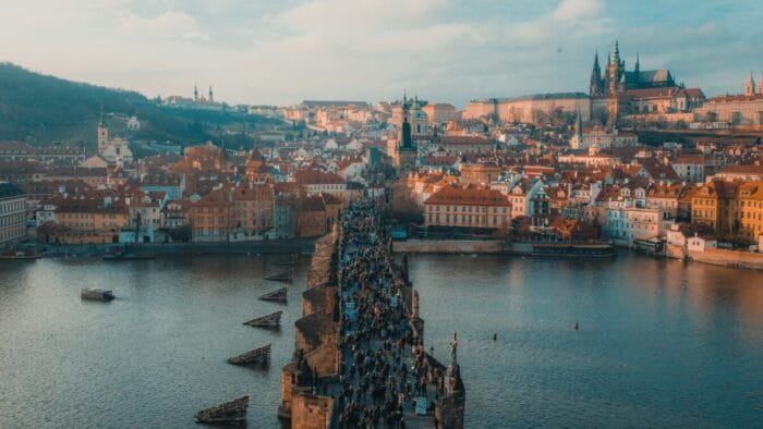 A multitude of people on pedestrian bridge over water in Prague with the skyline and a cathedral in the background