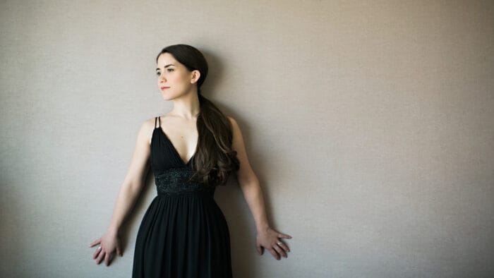 Joélle Harvey, in a black dress, poses against a cream colored wall