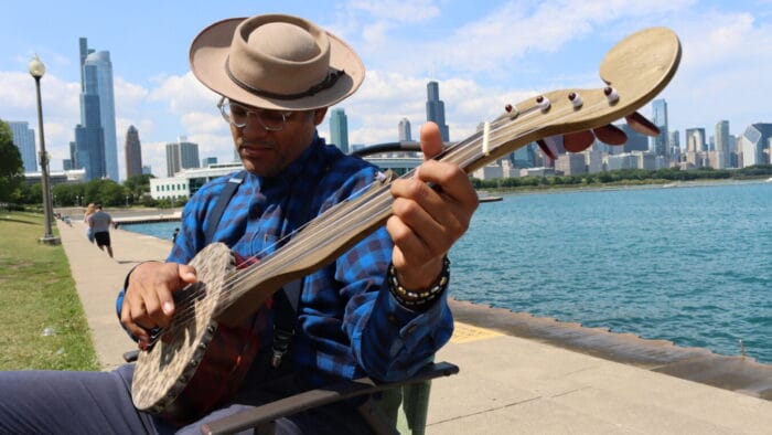 man in porkpie hat sits with banjo on lakeshore with Chicago skyline in background