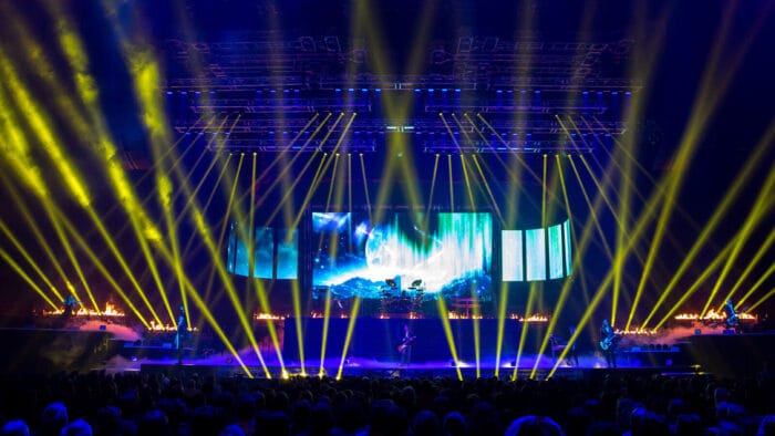 a blue stage lit up with yellow lights as a band performs to a packed audience