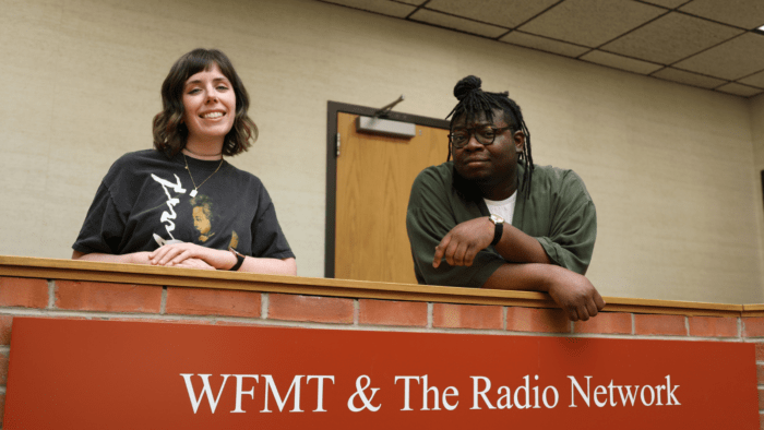Kristina and LaRob pose behind a brick wall and a sign that reads "WFMT & The Radio Network"