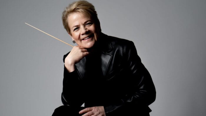 Marin Alsop sits cheerfully, wearing a black leather jacket and holding a conductors baton against a white backdrop