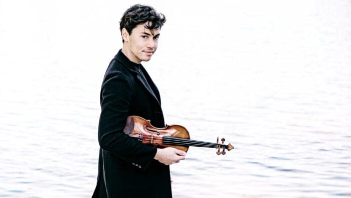 Benjamin Beilman, dressed in black, holds a violin while walking along the water