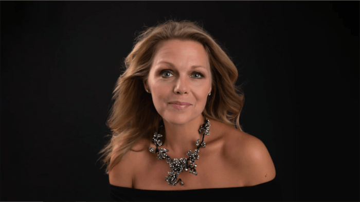 Miah Persson looking directly into camera in a strapless black dress with an ornate silver beaded necklace, dark background .