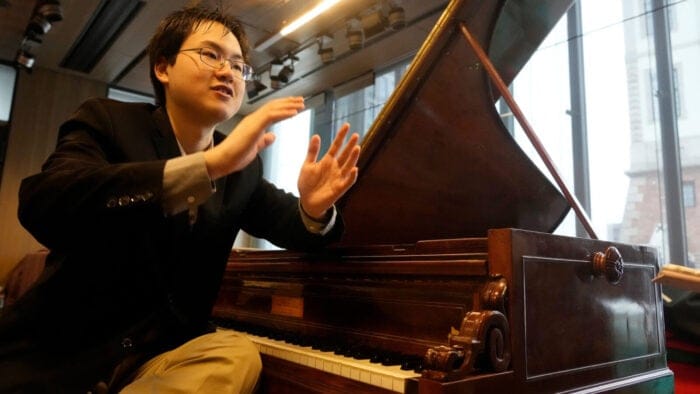 Pianist Eric Guo gestures excitedly sitting next to a historic keyboard