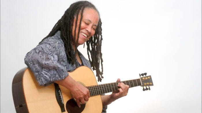 Black woman with long braids smiles and plays guitar