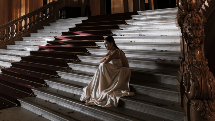 Hera Hyesang Park in cream colored goddess gown, sitting in the middle of a grand marble staircase framed by ornate baroque banisters.