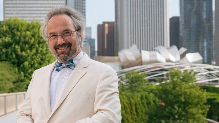 A color portrait of Carlos Kalmar, who poses in a white suit and blue patterned bowtie in front of the Chicago Skyline and Millennium Park's Pritzker Pavilion