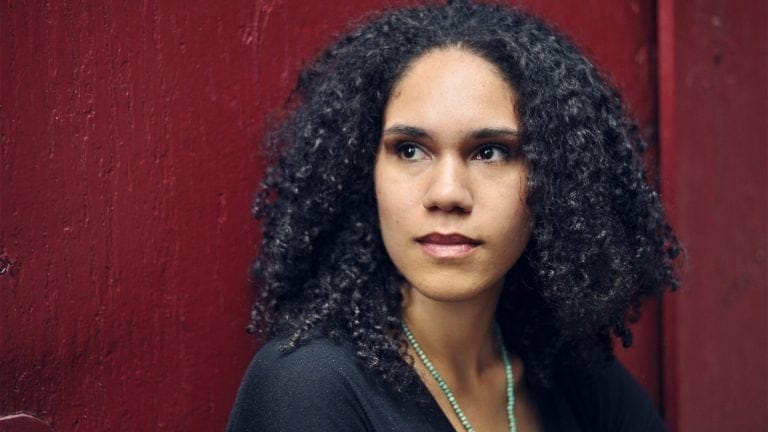 wearing a green necklace and a black sweater, Jessie Montgomery looks past the camera standing against a rich red wall