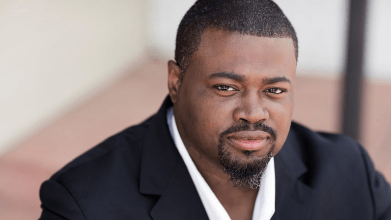 Russell Thomas headshot looking over right shoulder, black suit, pen collar white dress shirt