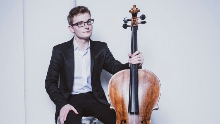 Alexander Hersh, wearing a dark suit and light shirt, poses with his cello