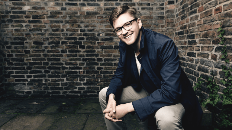 Samuel Hasselhorn portrait, in blue sportscoat and eyeglasses squatting in front of a brick wall