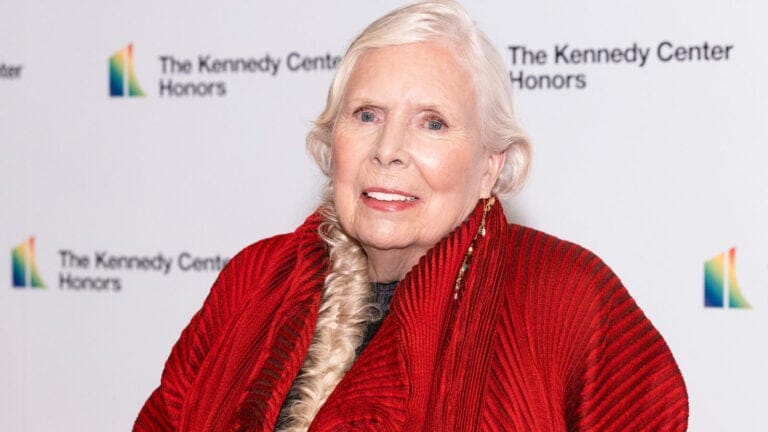 Joni Mitchell, dressed in red, smiles at the camera in front of a wall that says "The Kennedy Center Honors"