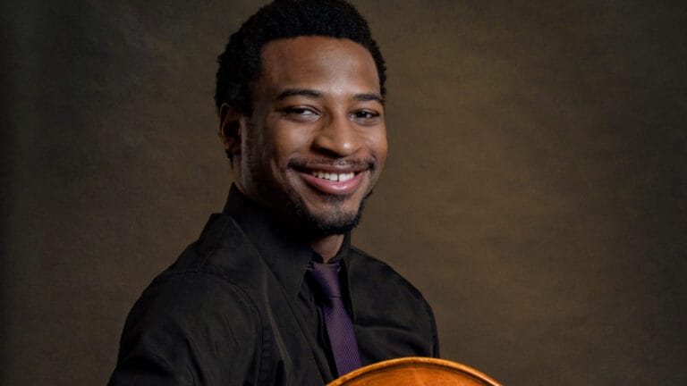 Xavier Foley, similing, poses with his double bass in front of a brown background