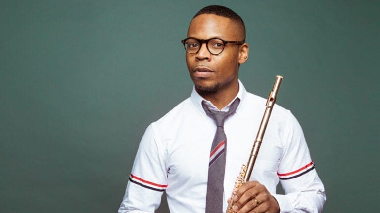 Brandon Patrick George, wearing a white shirt and grey striped tie, holds a flute in front of a green background
