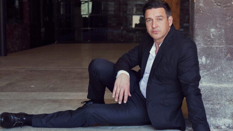 Color portrait of nathan Gunn wearing dark suit, white shirt, no tie, seated leaning against a brick wall