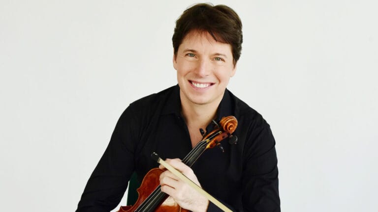Joshua Bell, smiling, sits in front of a white background with a violin and bow
