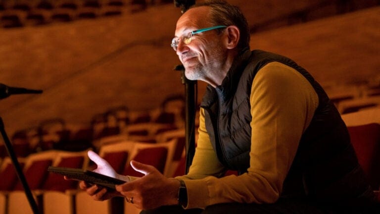Osvaldo Golijov holds a tablet and smiles, sitting in an empty auditorium