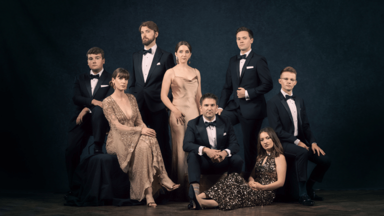 Portrait of VOCES8 in formal attire; three women in metallic-palette gowns; five gentlemen in black tie tuxedos; textured navy backdrop.