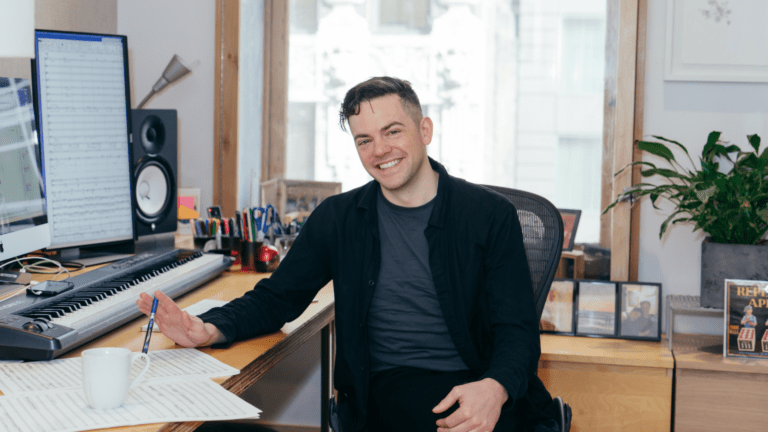 Nico Muhly in his composing studio, digital keyboard, sheet music, and large computer monitor on his desk, smiling, leaning on his right elbow; wearing black coat over grey-blue t-shirt.
