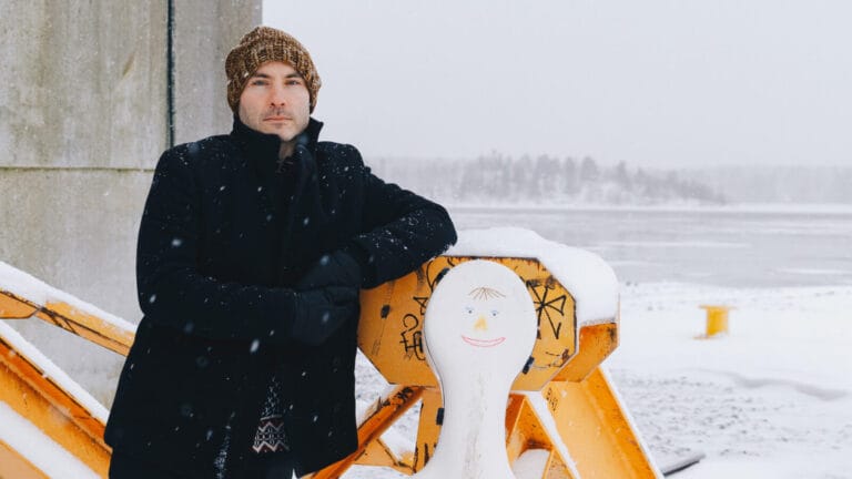Jakob Koranyi poses in front of a snowy backdrop with a while cello case bearing a smiley face