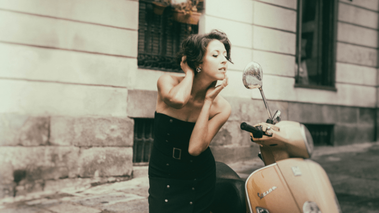 Lisette Oropesa in fitted strapless black dress with gold buttons, on the streets of Madrid, checking her makeup in the side mirror of a Vespa scooter.