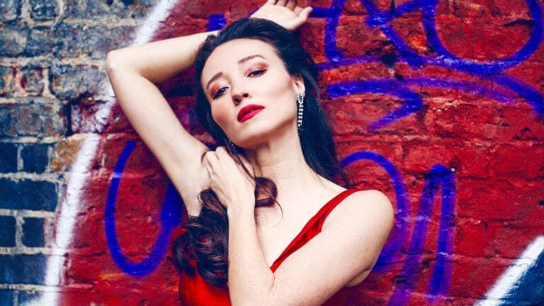 mezzo-soprano Fleur Barron poses in a red dress against a brick wall adorned with blue and red graffiti