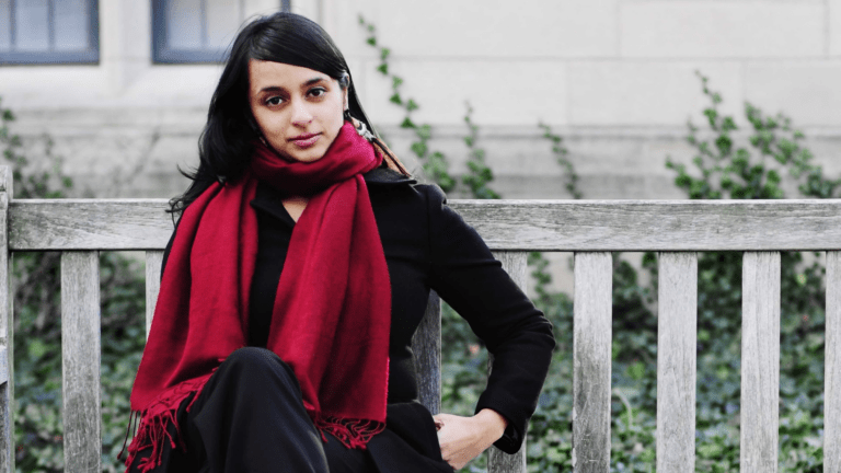 Reena Esmail in a black pantsuit with a generous, deep red scarf on a park bench, gray, natural background.