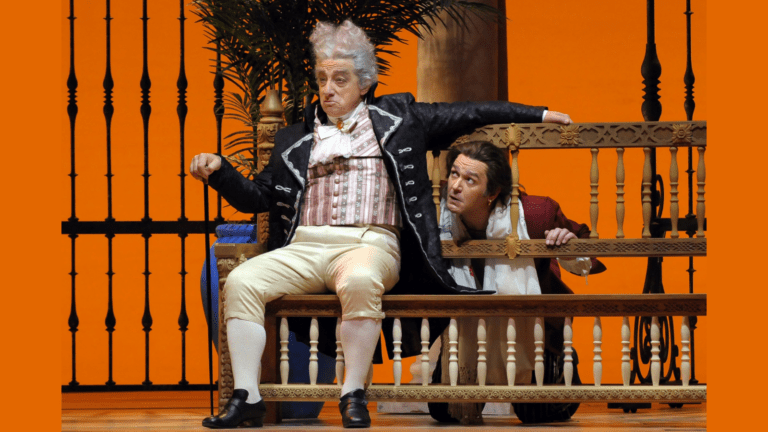 Alessandro Corbelli and Nathan Gunn in a scene from the Barber of Seville set in 18th century spain; Corbelli as Dr. Bartolo sitting on a bench looking disgruntled with Gunn as Figaro peeking mischievously at him from behind.