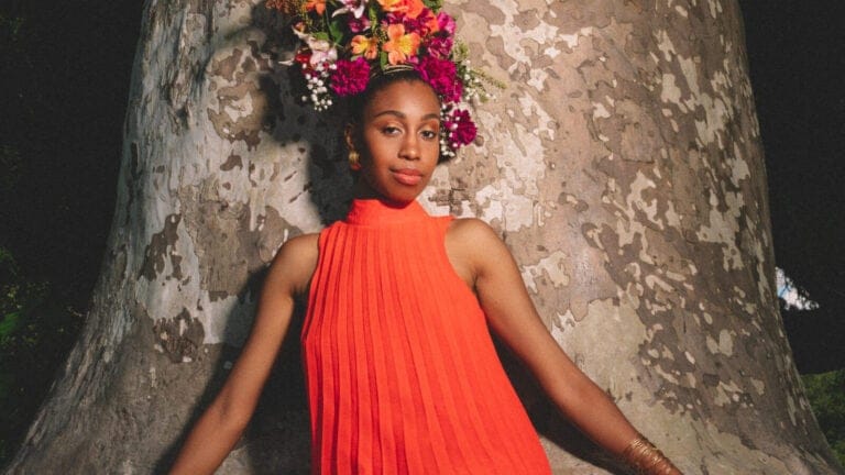 Jazzmeia Horn, wearing a flowery headdress and a bright orange-red dress, stands in front of a tree trunk