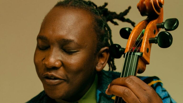 Abel Selacoe, in a vibrant blue suit and green shirt, is immersed in music as he plays the cello with eyes closed