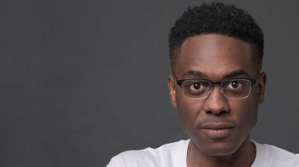 Headshot of Shawn Okpebholo, looking straight at camera wearing black half-frame glasses and white t-shirt.,