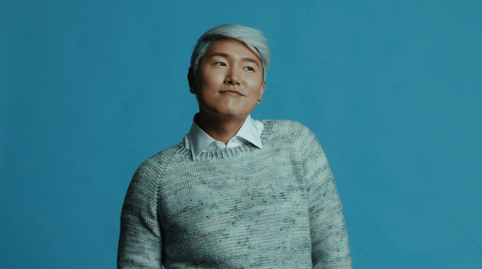 Kangmin Justin Kim with wry smile in hand-knit sweater, vivid aqua background