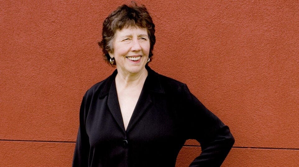 Joan Tower smiles, dressed in black, posing against a brick red stucco wall